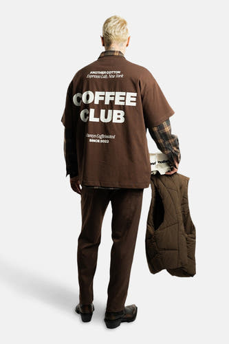 T-shirt marron à manches courtes d'ANOTHER COTTON LAB, avec une inscription sur le dos, porté avec un pantalon marron et une veste.