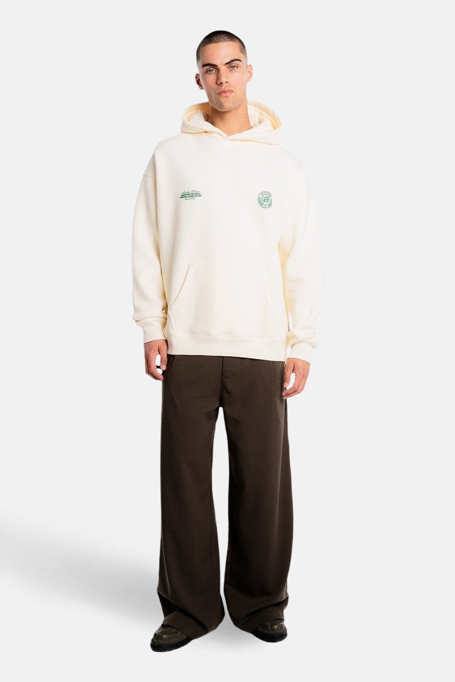 Pull à capuche écru de ANOTHER COTTON LAB, avec impressions de logo vertes et une poche kangourou, combiné avec un pantalon marron évasé et des chaussures noires.