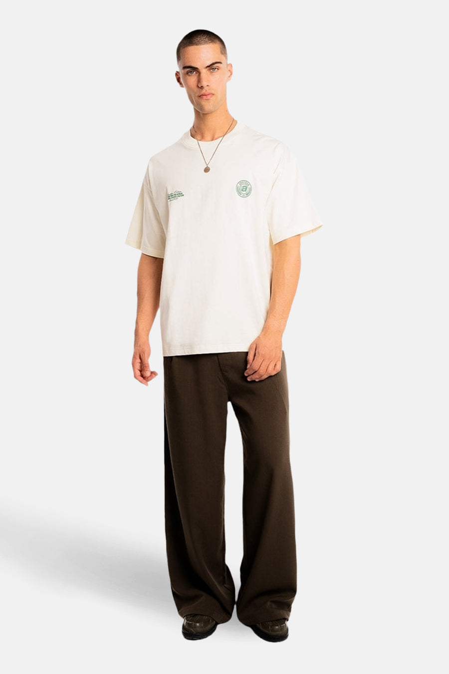 T-shirt écru à manches courtes de ANOTHER COTTON LAB, avec imprimé logo vert et combiné avec un pantalon marron et une chaîne dorée.