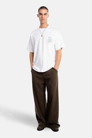 T-shirt blanc à manches courtes de ANOTHER COTTON LAB, avec un petit imprimé vert sur la poitrine et porté avec un pantalon marron et une chaîne.