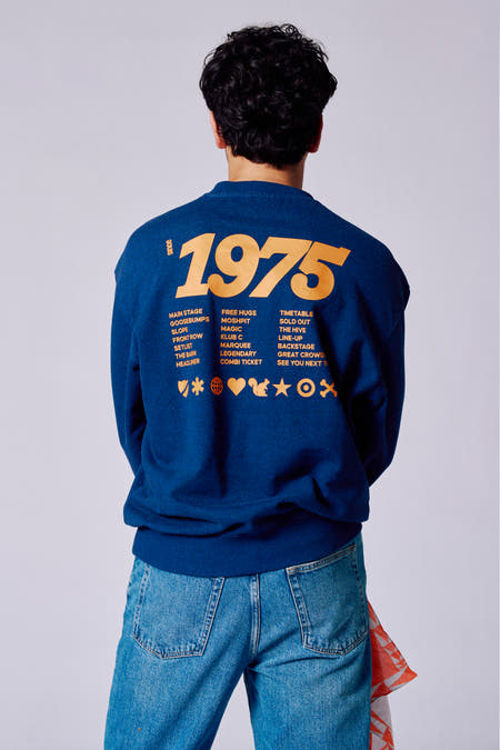 Pull-over bleu avec col rond de Rock Werchter 50, avec une impression jaune sur le dos et porté sur un pantalon de cuir.