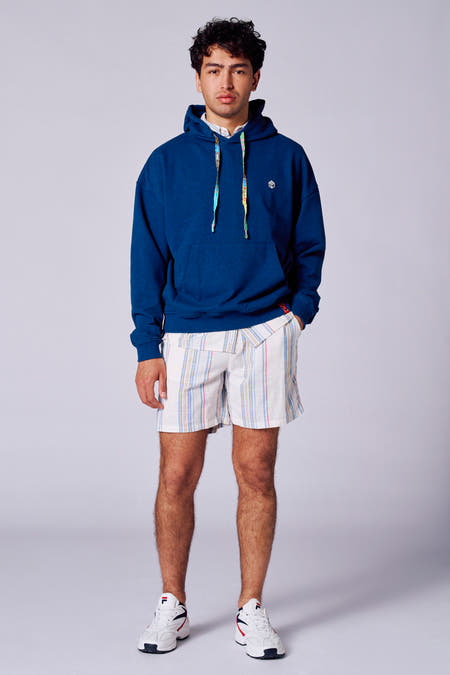 Blauwe sweater met kap van Rock Werchter 50, met trekkoorden en een logo, gecombineerd met gestreepte korte broek en witte sneakers.
