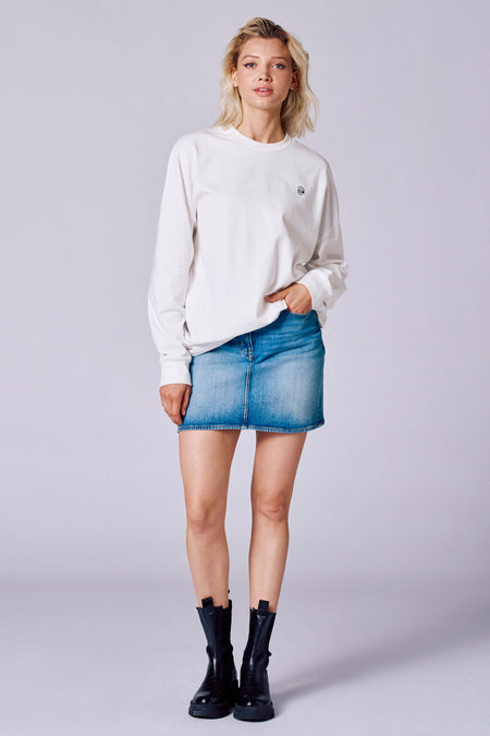 T-shirt blanc à manches longues de Rock Werchter 50, porté avec une jupe en denim et des bottes noires.