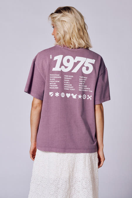 T-shirt violet à manches courtes de Rock Werchter 50, porté avec une jupe blanche à volants, un sac à dos et des bottes noires.