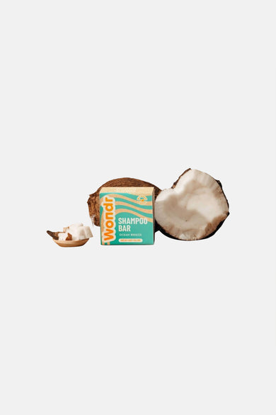 Barre de shampoing dans une boîte turquoise avec motif ondulé beige de WONDR CARE, entre des demi-noix de coco et un petit bol en bois avec des morceaux de noix de coco.
