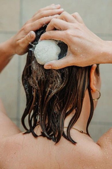 Gel douche solide blanc de WONDR CARE mousse sur les cheveux foncés mouillés d'une personne sous la douche, avec une boucle d'oreille dorée.
