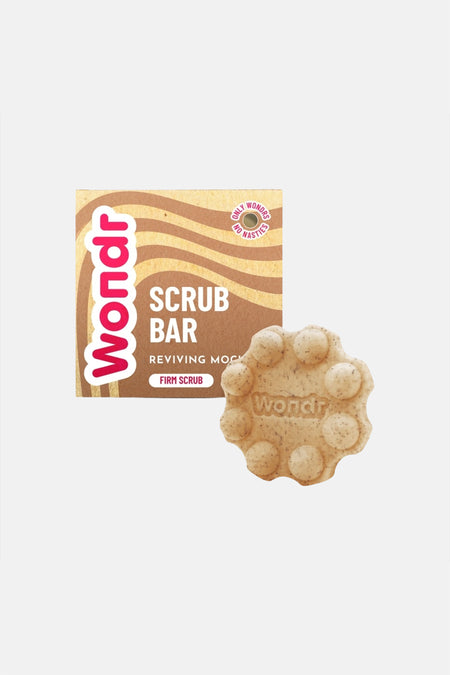 Une barre de gel douche beige clair avec des particules exfoliantes de WONDR CARE, ronde avec des picots et le texte 'ndr', moussant sur un bras sous la douche.