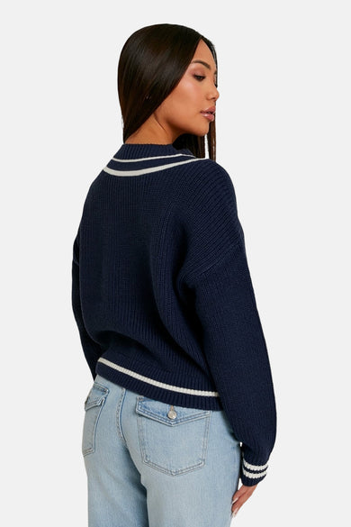 Pull bleu à col en V de TALLY WEIJL, avec des rayures blanches sur le col et les manches, porté avec un jean.