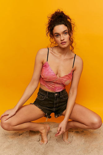 Vrouw in roze top, zwarte shorts, gehurkt op zand, gele achtergrond.