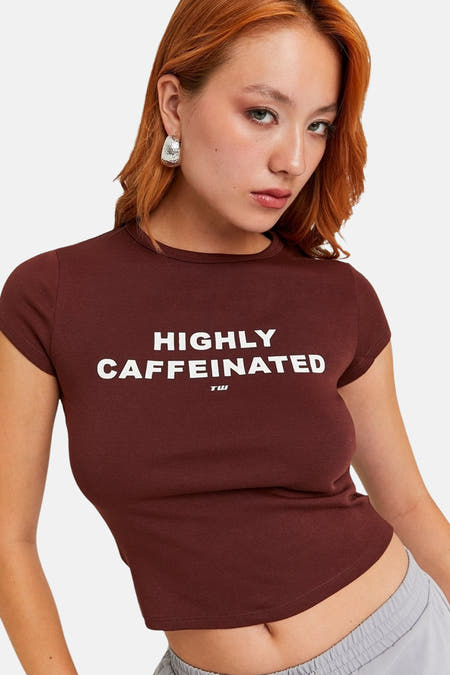 T-shirt marron à manches courtes de TALLY WEIJL, avec le texte "HIGHLY CAFFEINATED" et porté avec des boucles d'oreilles en argent.
