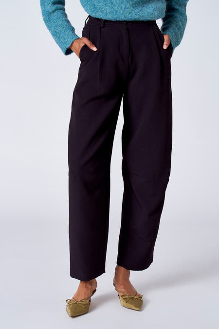 Pantalon de costume noir de SUNSET STUDIO, avec des poches à l'arrière et une main sur le pantalon.