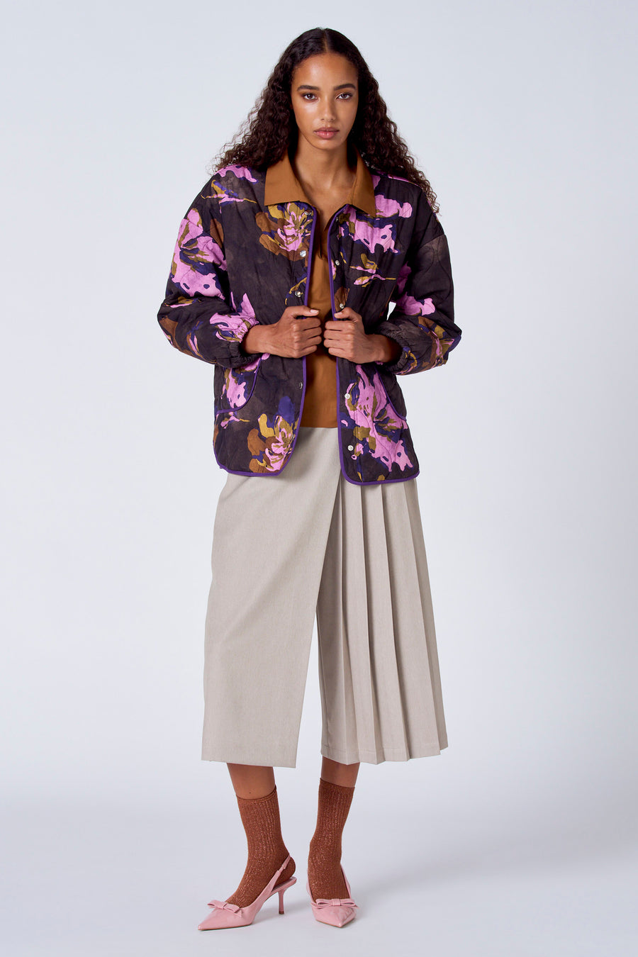 Chemisier marron à manches longues de SUNSET STUDIO, porté sous une veste fleurie avec un pantalon culotte beige, des chaussettes marron et des chaussures roses.