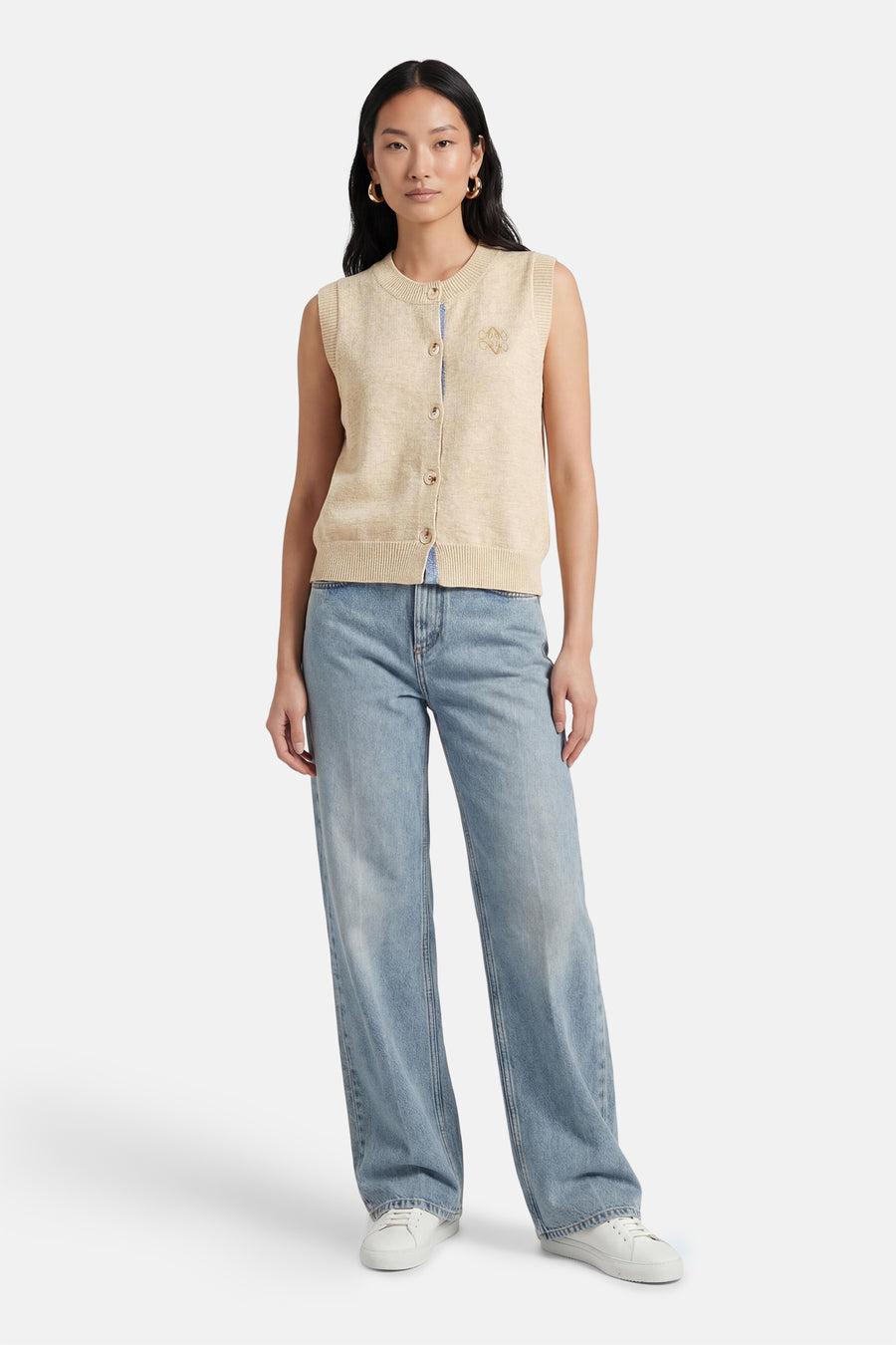 Pull beige ADORE, sans manches, avec fermeture à boutons et écusson, combiné avec un jean bleu et des baskets blanches.