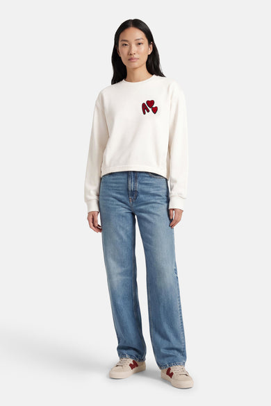 Pull blanc à col rond ADORE, orné d'une lettre rouge et de cœurs, porté avec un jean bleu et des baskets crème.