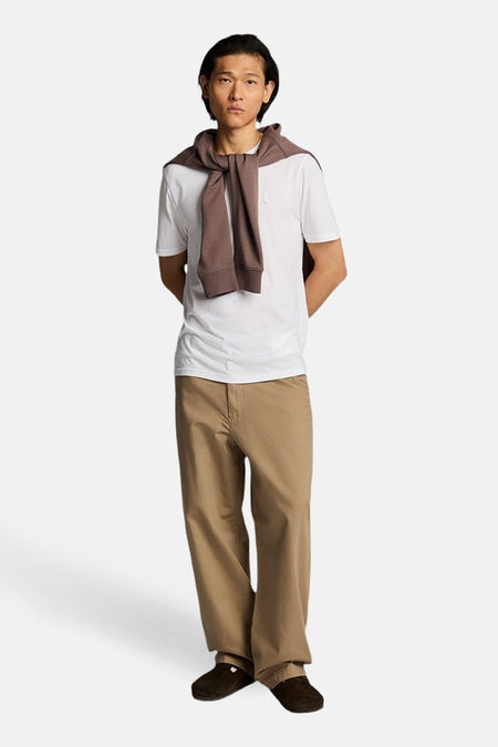 T-shirt blanc à manches courtes de LYLE & SCOTT porté par un homme debout avec un pull marron sur les épaules et un pantalon beige.