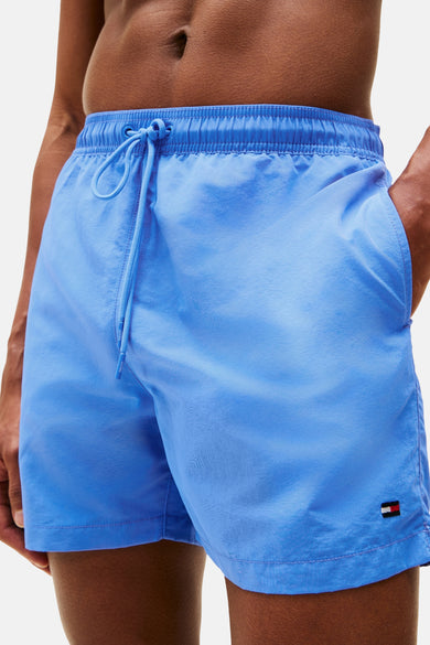 Short de bain bleu Tommy Hilfiger, porté avec une chemise blanche en lin ouverte et un sac en toile sur l'épaule.