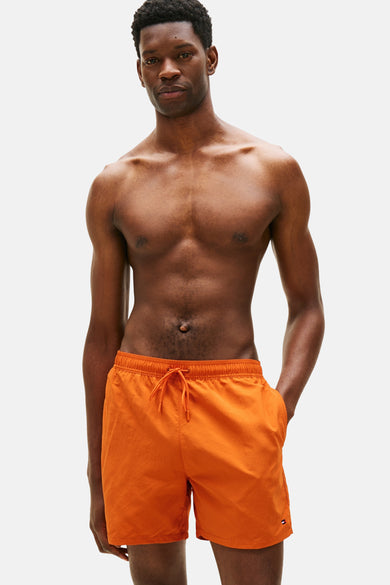 Short de bain orange Tommy Hilfiger, porté par un homme les mains dans les poches, sur fond blanc.