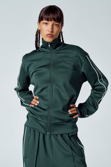Veste vert foncée Un Unwind avec deseses blanches, portée par un modèle aux cheveux tressés et aux boucles d'oreilles dorées.