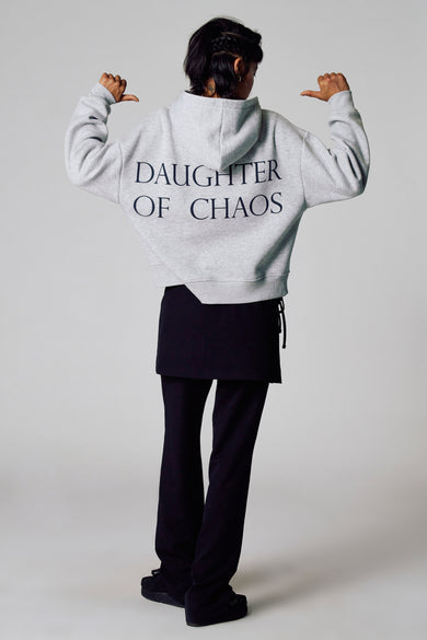 Pull-over gris avec capuche de Unwind, avec le texte "DAUGHTER OF CHAOS" sur le dos, porté avec une jupe noire et un pantalon.