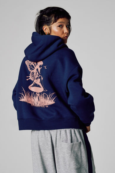 Pull-over bleu avec capuche Unwind, imprimé de fée rose sur le dos, porté sur un short de jogging gris.