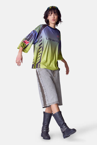 T-shirt vert avec manches courtes d'Unwind, avec dégradé de couleurs et motifs graphiques, porté avec des shorts en denim et des bottes noires.