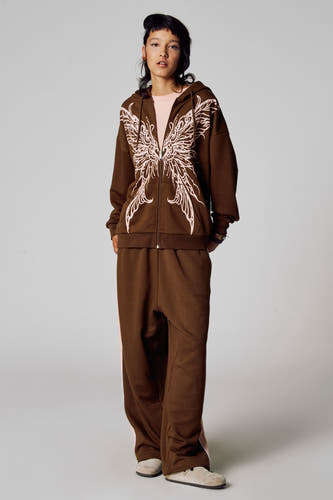 Pull-over marron d'Unwind, avec fermeture éclair et motif de aes roses, associé à un short de jogging coordonné et des chaussures beige.