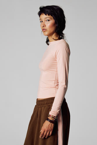 T-shirt rose à manches longues d'Unwind, avec des bijoux autour du poignet et une jupe brune, en vue latérale.