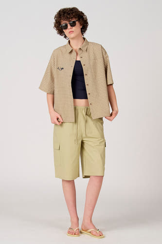 Chemise beige à manches courtes d'Unwind, à motif à carreaux et avec un logo sur la poitrine, portée sur un haut foncé et un short kaki.