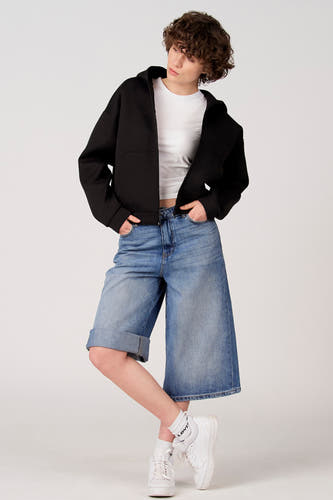 Gilet pull noir d'Unwind, porté sur une chemise blanche, avec un bermuda en jean ample et des baskets blanches.