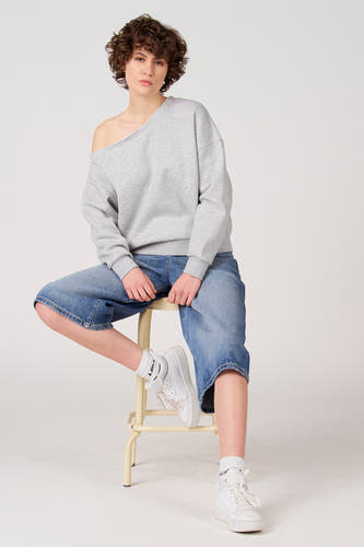 Pull gris Unwind, porté avec les épaules dénudées, combiné avec un jean ample et des baskets blanches sur un tabouret.