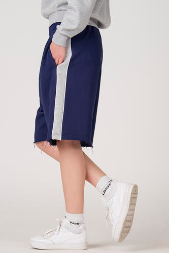 Short bleu d'Unwind avec rayure grise sur le côté et bord effiloché, porté avec des chaussettes et des chaussures blanches.