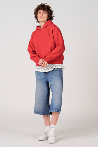Pull rouge à capuche Unwind, avec poche kangourou et haut en dentelle blanche visible, combiné avec un pantacourt en jean et des baskets blanches.