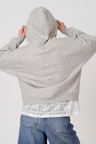 Sweat à capuche gris Unwind, avec cordon de serrage, une poche kangourou et portable sur une blouse blanche en dentelle et un jean bleu.