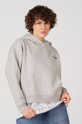 Grijze sweater met kap van Unwind, met trekkoord, een buidelzak en draagbaar over een witte kanten blouse en blauwe jeans.
