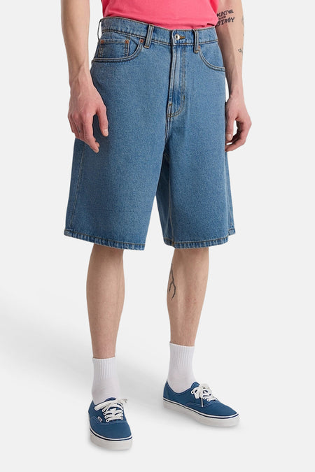 Short en jean bleu clair de VANS "OFF THE WALL" avec poches arrière plaquées, porté par une personne tatouée avec des chaussures en toile bleue.