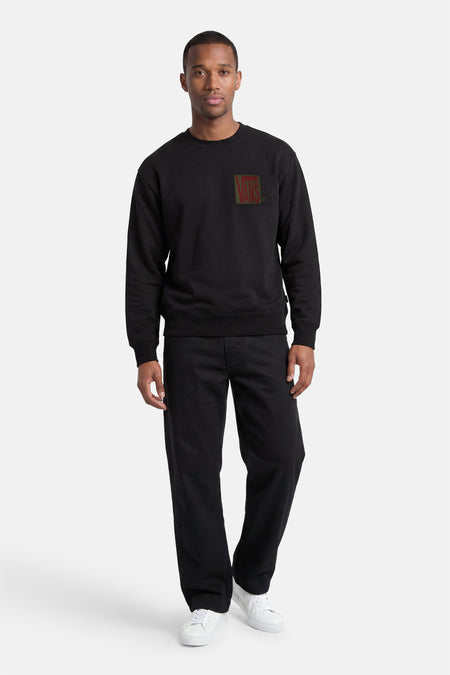 Pull noir à col rond VANS "OFF THE WALL" avec logo VANS rouge et vert sur la poitrine, porté avec un pantalon noir et des baskets blanches.
