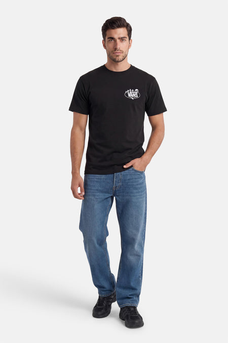 T-shirt noir de VANS avec un logo ovale blanc sur la poitrine, porté par un homme avec un jean bleu et des baskets noires.