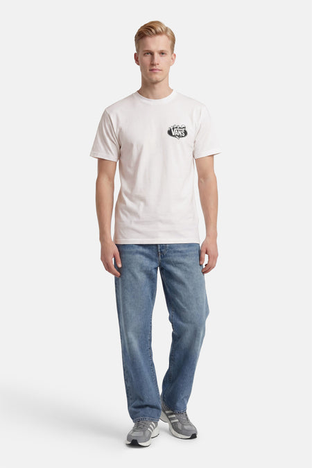 T-shirt blanc VANS "OFF THE WALL" avec logo noir sur la poitrine, porté par un homme avec un jean bleu et des baskets grises.