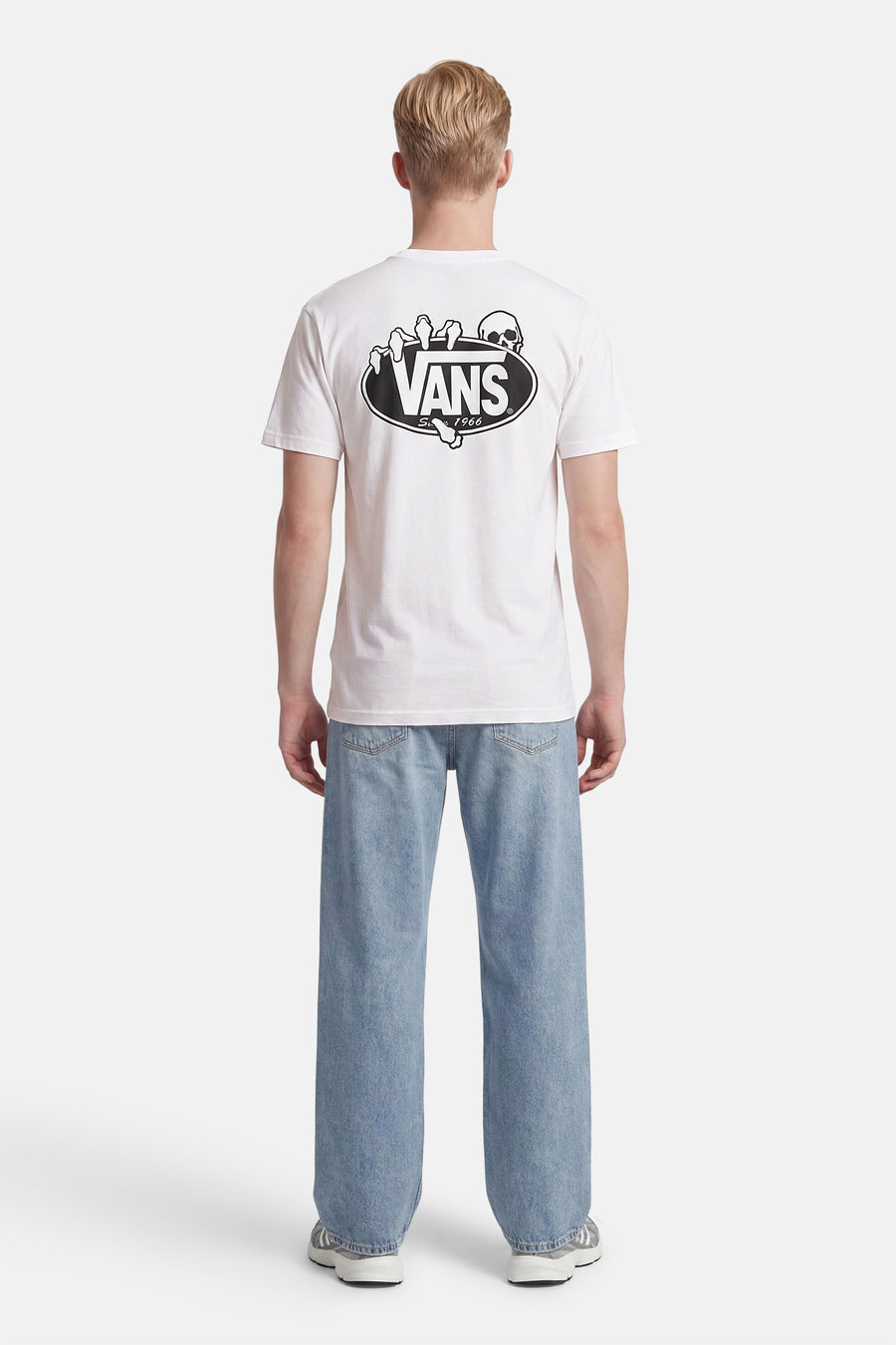 T-shirt blanc VANS avec imprimé tête de mort et main dans le dos, porté avec un jean bleu clair et des baskets grises.