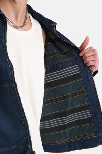 Veste en jean bleu foncé VANS "OFF THE WALL", avec une doublure à carreaux et portée sur une chemise blanche avec une chaîne argentée.