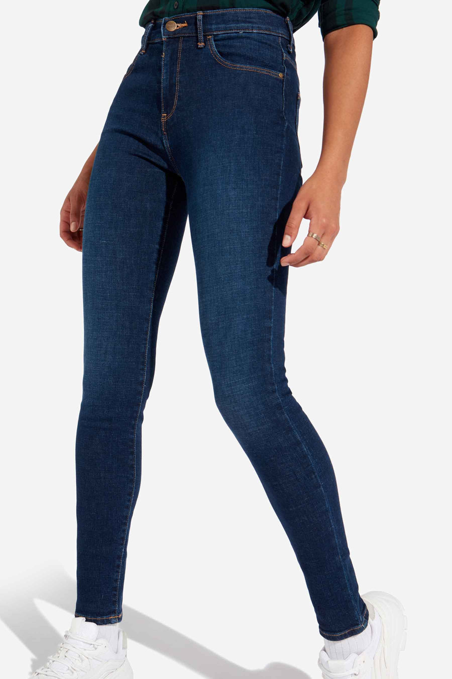 Jean skinny bleu marine de Wrangler, avec surpiqûres dorées et porté avec des baskets blanches.