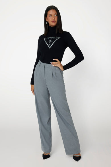 Col roulé noir Guess® avec logo triangulaire Guess scintillant, porté par un mannequin avec un pantalon plissé gris et des talons aiguilles noirs.