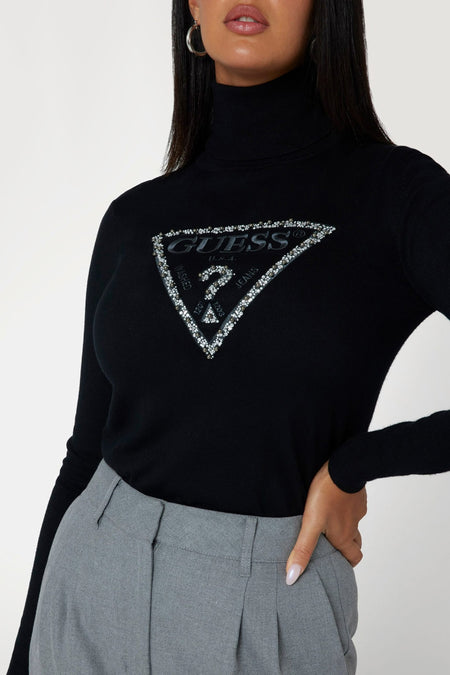 Pull noir à col roulé Guess® avec logo strass, porté avec un pantalon gris.