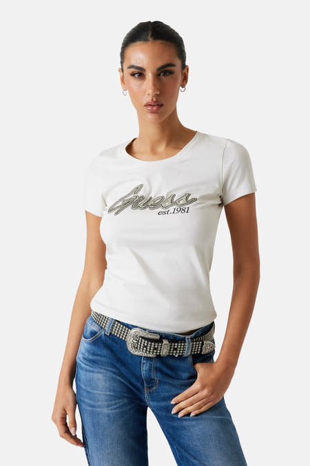 T-shirt blanc à manches courtes de Guess, avec des paillettes et le texte "est. 1981", porté avec un jean et une ceinture.