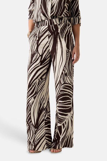 Pantalon écru à imprimé marron de Guess® avec jambes larges, un motif de feuilles abstrait, un chemisier assorti et des sandales dorées.