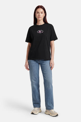T-shirt noir à manches courtes de Guess, avec un imprimé logo rose, combiné avec un jean bleu et des baskets couleur crème.