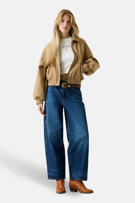 Model in een beige jas, wit T-shirt, blauwe wijde pijpen jeans en bruine laarzen.