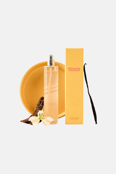 Parfum jaune de WONDR CARE dans une bouteille transparente avec une boîte jaune, une gousse de vanille, une fleur et de l'écorce d'arbre sur un plat jaune.