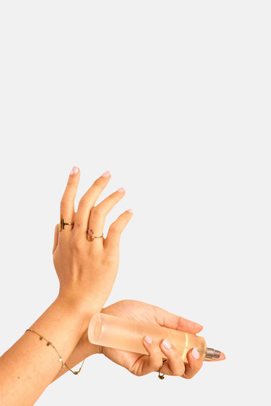 Flacon de parfum transparente avec un liquide légèrement orangé de WONDR CARE, tenue par des mains avec des bagues dorées et un bracelet.