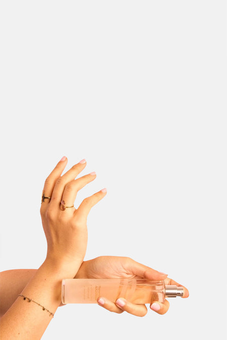Parfum rose clair dans un flacon en verre de WONDR CARE, tenu horizontalement par des mains avec des bagues et un bracelet.